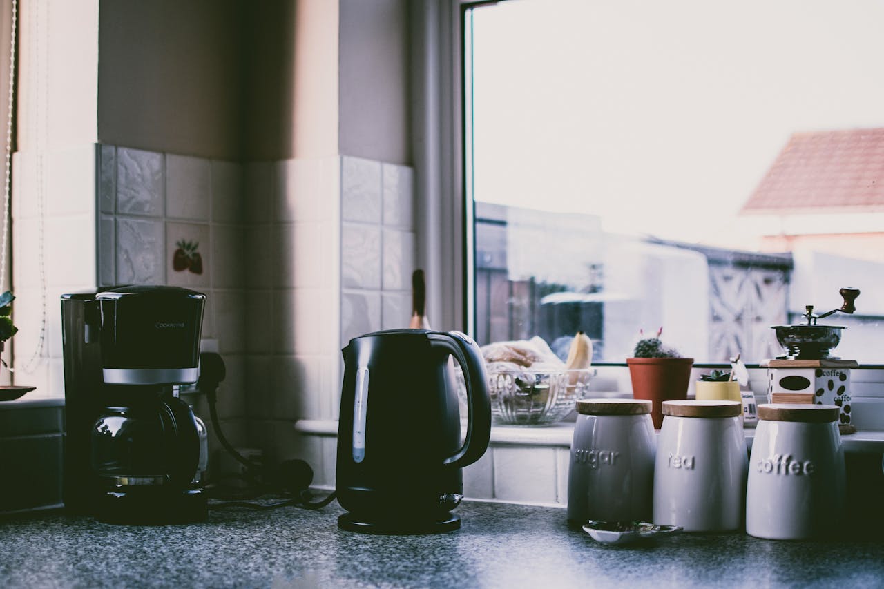 Services Cozy kitchen scene with coffee maker, kettle, and jars on a sunny counter.